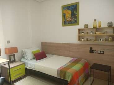 Este acogedor dormitorio cuenta con una cama individual con almohadas de colores y una manta, una mesilla de noche, estanterías de pared con decoración, una lámpara y modernas obras de arte abstractas.