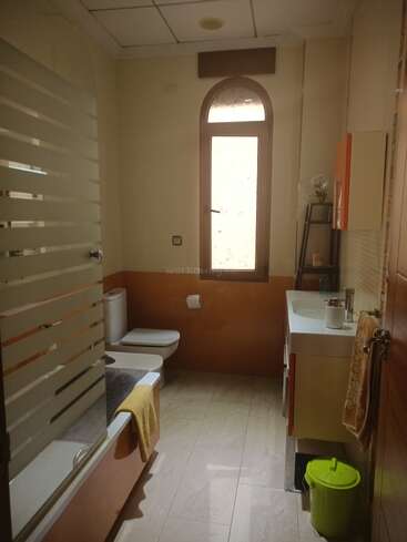 Este cuarto de baño tiene bañera con mampara de cristal, inodoro, lavabo, ventana arqueada, toallas, estantería con decoración y una papelera verde en el suelo de baldosas.