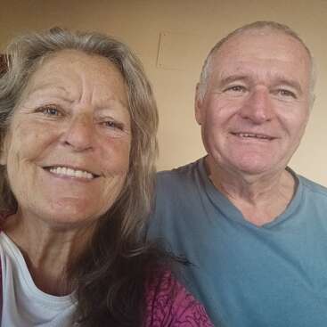 Una anciana y un anciano sonrientes se toman un alegre selfie juntos en un interior. Aparecen felices y relajados, vestidos con ropa informal, con un fondo liso detrás de ellos.