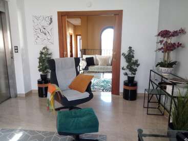 Modernes Wohnzimmer mit einem bequemen Stuhl, einer Fußbank, bunten Kissen und einer Decke. Pflanzen, Blumen und eine minimalistische Einrichtung schaffen eine helle, einladende und stilvolle Atmosphäre.