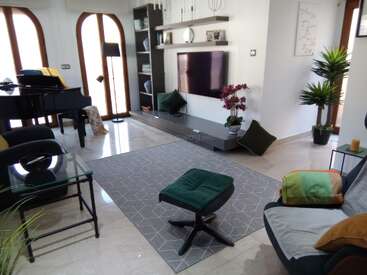 In diesem modernen Wohnzimmer sorgen ein Flügel, ein an der Wand montierter Fernseher, ein grauer Teppich, grüne Polstermöbel, Pflanzen und große Bogenfenster für einen hellen, einladenden Raum.