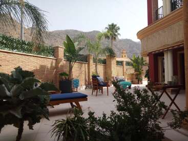 Eine sonnige Terrasse mit Liegestühlen, tropischen Pflanzen und Palmen ist von einer Backsteinmauer umgeben. Dahinter sind die Berge und ein Kirchturm zu sehen.