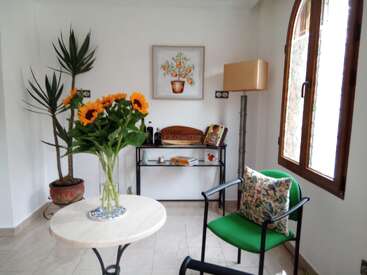 Ein gemütliches, sonnendurchflutetes Zimmer mit einem grünen Stuhl, Blumenkissen, einer Vase mit Sonnenblumen, einer Topfpflanze, gerahmter Blumenkunst, Bücherregalen und einem großen Fenster.