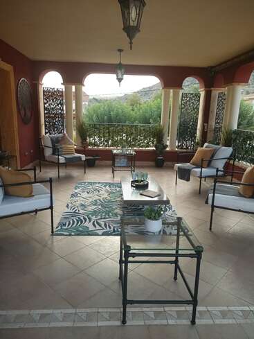 Überdachte Terrasse mit bequemen Sitzgelegenheiten, gemustertem Teppich, Glastischen, dekorativen Kissen, hängenden Laternen, Topfpflanzen, malerischem Naturblick, roten Wänden und verschlungenen Metallpaneelen.