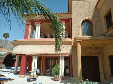 Dieses Bild zeigt eine Villa im mediterranen Stil mit roten und beigen Tönen, gewölbten Fenstern, einem Balkon, Palmen, Terrassenmöbeln und einem sonnigen Außenbereich.