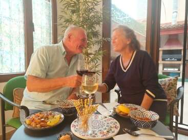 Una pareja mayor se sienta sonriente y brinda con vino en una acogedora mesa, disfrutando de una comida juntos en el interior, rodeados de vajilla de colores y una cálida luz natural.