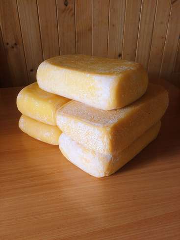 A imagem apresenta uma pilha de quatro blocos retangulares de queijo com uma tonalidade amarelo-alaranjada e casca branca, colocados em uma mesa de madeira contra um fundo de parede de madeira.