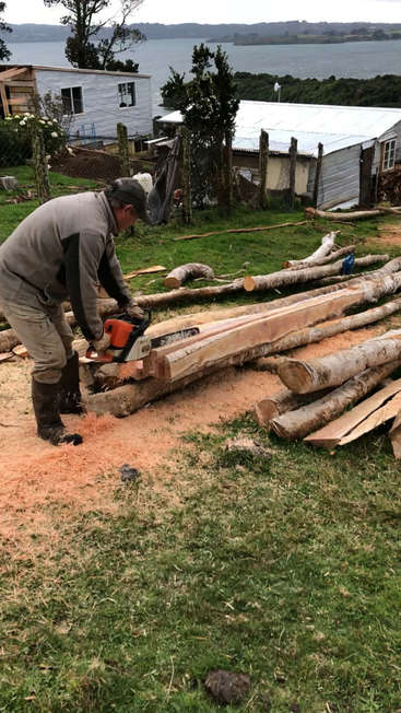 Um homem está usando uma motosserra para cortar um tronco grande em uma área gramada perto de um corpo d'água e de alguns edifícios.