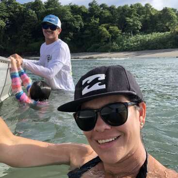 A woman takes a selfie in the water, smiling, wearing sunglasses and a cap. Behind her, a man helps a child near a boat by the beach.
