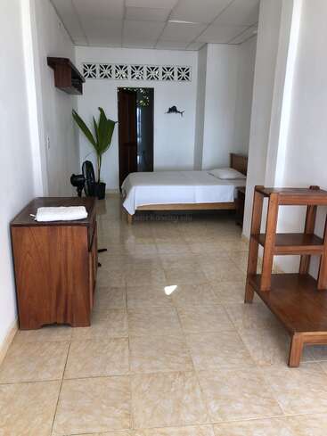 This image shows a simple, clean room with tiled floor, a single bed, wooden furniture, a potted plant, ceiling tiles, and a fish wall decoration.