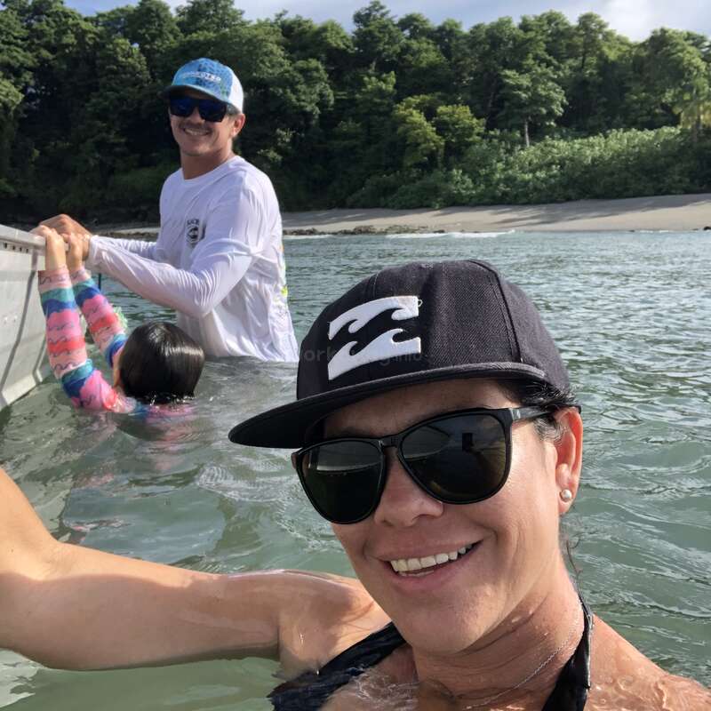 A woman takes a selfie in the water, smiling, wearing sunglasses and a cap. Behind her, a man helps a child near a boat by the beach.
