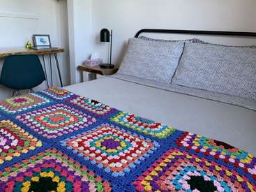 Un dormitorio acogedor con una cama con cojines estampados y una colorida manta de ganchillo. Hay un escritorio de madera, una silla verde, una lámpara, pañuelos y cuadros de arte.