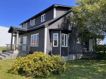 Una casa de dos plantas de color gris oscuro con adornos blancos se encuentra bajo un cielo azul despejado. Tiene un porche, varias ventanas, hierba verde, arbustos y árboles.