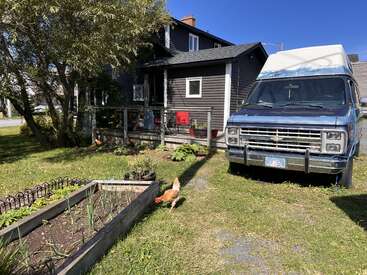 Una casa gris con un porche delantero, sillas rojas, gallinas deambulando por el patio, un huerto, una gran furgoneta aparcada y un exuberante entorno verde.