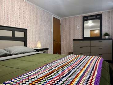 Esta habitación cuenta con papel pintado floral, una cama con un edredón verde, dos almohadas grises, una manta de ganchillo de colores, una cómoda, un espejo, una lámpara y una planta.