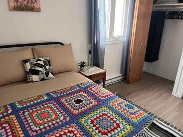 Una acogedora habitación cuenta con una cama con ropa de cama neutra, una colorida manta de ganchillo, un cojín geométrico, una mesita, un armario abierto y una ventana luminosa.