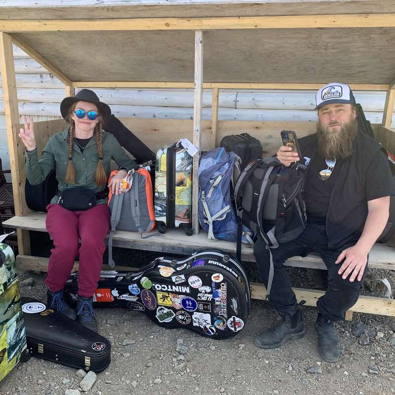 La imagen muestra a un hombre y una mujer sentados en un banco de madera, rodeados de equipaje y equipo musical, con una rústica pared de cabaña de madera al fondo.