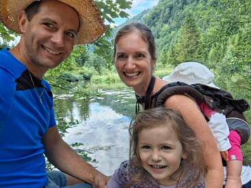 A smiling family of four poses by a tranquil lake surrounded by lush green trees and mountains. The atmosphere is joyful, outdoorsy, and full of nature.