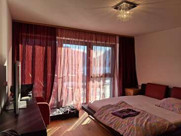 A cozy hotel room with a large bed, television, and red curtains letting in sunlight from a balcony. Warm lighting and modern, comfortable decor throughout.