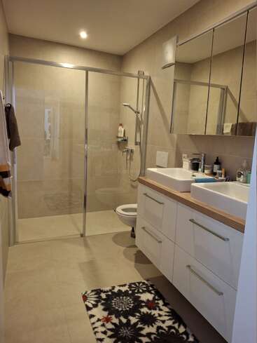 This modern bathroom has beige tiles, a glass shower enclosure, double sinks, large mirrors, white cabinets, and a patterned rug, creating a tidy, inviting atmosphere.