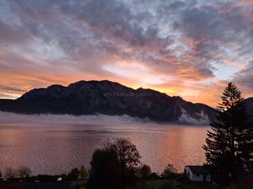 Un lac serein reflète le ciel spectaculaire du coucher de soleil, les silhouettes des montagnes et les nuages qui défilent. Les arbres et la maison au premier plan créent un paysage paisible et pittoresque.