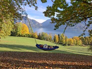 Une balancelle suspendue à un arbre surplombe un lac paisible et les montagnes. Deux enfants casqués se détendent, entourés d'un feuillage automnal luxuriant et d'un paysage aux couleurs vives.