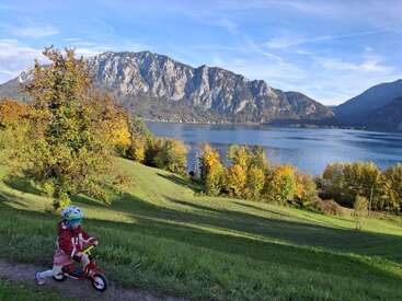 Un jeune enfant roule à vélo sur une colline verdoyante, avec des arbres automnaux, un lac calme et des montagnes majestueuses sous un ciel bleu éclatant.