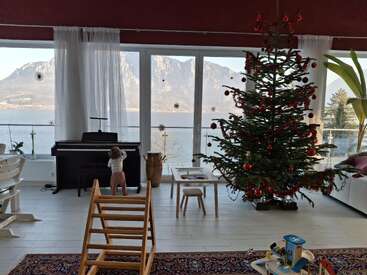Un salon confortable comprend un sapin de Noël décoré, un piano, des jouets et un enfant, avec de grandes fenêtres donnant sur de magnifiques montagnes et un lac à l'extérieur.