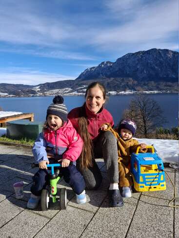 A smiling woman and two children enjoy a sunny winter day outdoors by a lake, with mountains in the background. The children play with toys.