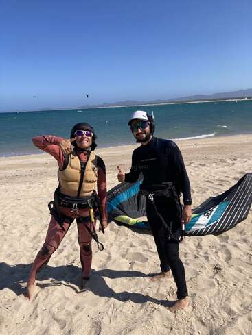 Zwei Menschen in Neoprenanzügen und Helmen stehen an einem Sandstrand, lächeln und zeigen die Daumen nach oben, während im Hintergrund die Kitesurfausrüstung und das Meer zu sehen sind.