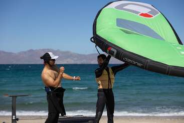 Zwei Menschen stehen am Strand, einer ohne Hemd und gibt Anweisungen, während der andere einen großen grünen Ozonflügel hält und sich auf das Windsurfen oder Foilen vorbereitet.