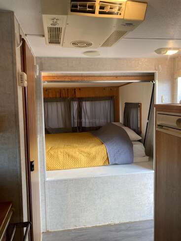 Dieses Bild zeigt das Innere eines Wohnmobils oder Wohnwagens mit einem gemütlichen Bett, einer gelben Decke, grauen Kissen, einer Holzverkleidung, einer Klimaanlage und kleinen Fenstern mit Vorhängen.