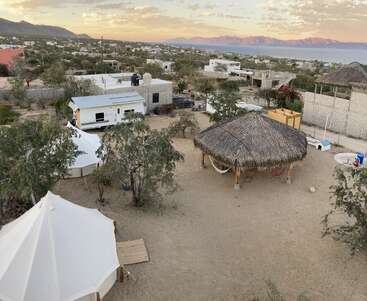 Ein sandiger Hof mit Bäumen, Zelten und einer großen Palapa liegt zwischen Häusern. Im Hintergrund sind die Berge und das Meer unter einem wolkenverhangenen Himmel zu sehen.