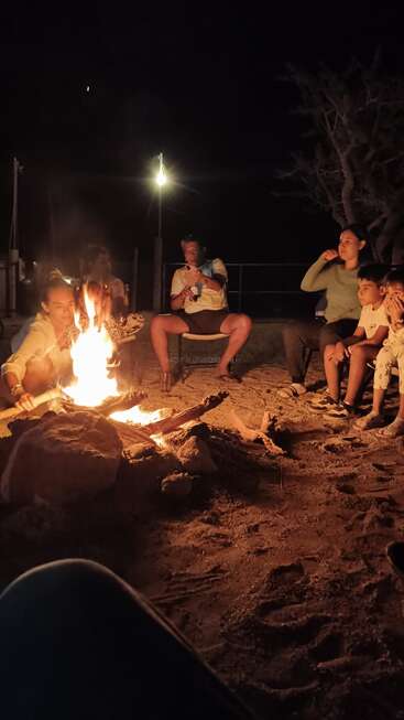 Eine Gruppe von Menschen, darunter auch Kinder, sitzt nachts um ein Lagerfeuer, genießt die warme Glut und genießt einen friedlichen, gemütlichen Moment im Freien.