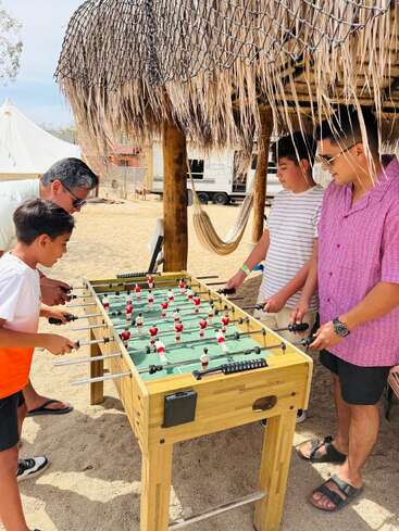 Vier Personen spielen unter einer strohgedeckten Hütte an einem Sandstrand Tischfußball. Sie scheinen konzentriert zu sein und genießen ein freundschaftliches Spiel im Freien.