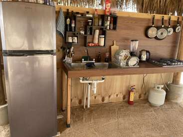 Dieses Bild zeigt eine Outdoorküche mit Kühlschrank, Spüle, Regalen mit Zutaten, Utensilien, Geschirrständer, Mixer, Wasserkocher, Herd, Pfannen und einer Gasflasche.