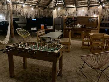 Diese gemütliche Außenküche ist mit einem Strohdach, Lichterketten, Holzmöbeln, einem Kickertisch, einer Hängematte und einem Kühlschrank ausgestattet und schafft eine rustikale, einladende Atmosphäre für Versammlungen.