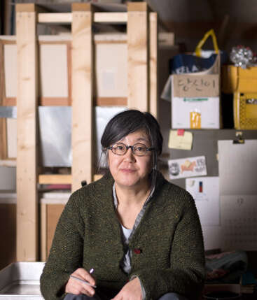 Une femme à lunettes et aux cheveux gris est assise à l'intérieur, un stylo à la main. Elle porte un pull-over vert. L'arrière-plan est encombré de boîtes, de papiers et de notes.