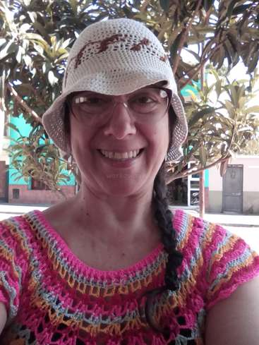 A imagem mostra uma mulher usando um chapéu de crochê branco e uma blusa de crochê rosa, amarela e azul, em pé em frente a árvores e edifícios.