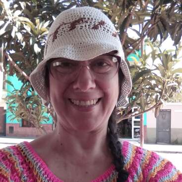 The image shows a woman wearing a white crocheted hat and a pink, yellow, and blue crocheted top, standing in front of trees and buildings.