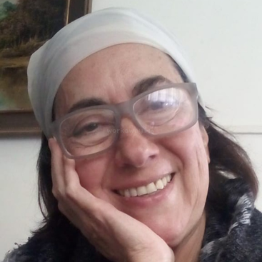 Uma mulher sorridente, usando óculos grandes e um lenço branco na cabeça, apoia o rosto na mão. Ela parece relaxada e alegre, com um quadro ao fundo.