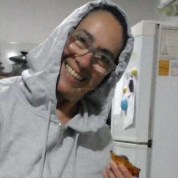 Uma pessoa sorridente, de óculos e moletom cinza com capuz, está dentro de casa, segurando um pedaço de comida. Ao fundo, dá pra ver uma geladeira decorada com ímãs e papéis.