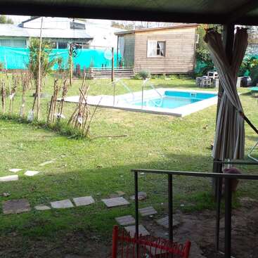 Una terraza cubierta con una silla roja da a un jardín trasero con césped, una piscina, una casita de madera, unos pasos de piedra y una valla, creando un ambiente relajante.
