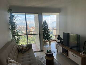 Im gemütlichen Wohnzimmer mit Sofa, Fernseher und Couchtisch steht ein geschmückter Weihnachtsbaum neben großen Fenstern mit Blick auf die Stadt und die Berge in der Ferne.