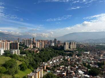 Dieses Bild zeigt eine lebendige Stadtlandschaft mit hohen Gebäuden, umgeben von üppigem Grün, unter einem blauen Himmel mit vereinzelten Wolken und fernen Bergen im Hintergrund.