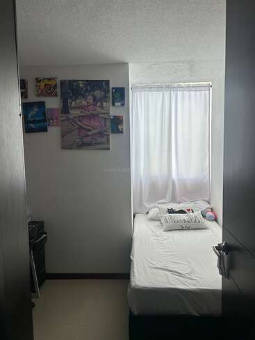 Ein kleines, gemütliches Schlafzimmer mit einem Einzelbett, weißem Bettzeug und Cartoon-Kissen. Ein Fenster mit weißen Vorhängen. Die Wand ist mit Familien- und Cartoon-Fotos dekoriert.