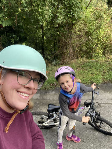 A woman and a young girl wearing helmets smile for a selfie outdoors. The girl stands next to her bicycle on a paved path surrounded by greenery.