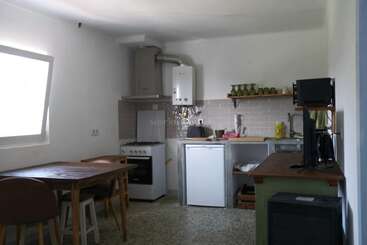 This image shows a small, simple kitchen with a dining table, chairs, stove, fridge, sink, television, microwave, and open shelves holding green cups.