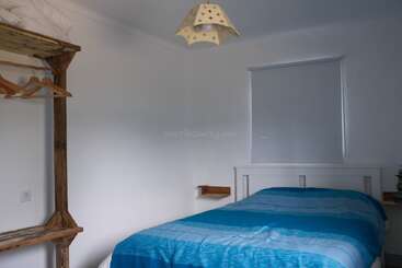 A simple bedroom with a blue bedspread, wooden open wardrobe with hangers, white walls, a small window with a blind, and a decorative ceiling lamp.