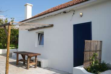 A small white house with a red-tiled roof features a wooden pergola, rustic picnic table, blue door, garden plants, and a bright, sunny atmosphere.